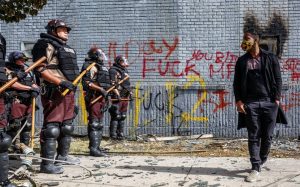 A man facing a line of riot cops in Minneapolis during George Floyd Uprising, behind is a wall with anti-police graffiti