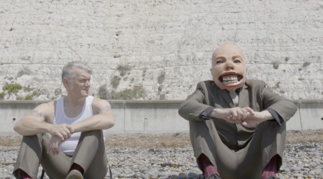 Dunstan Bruce sitting by a wall with someone wearing the baby face from the Tubthumper album cover and cartoon hands from “Never Mind The Ballots” album cover