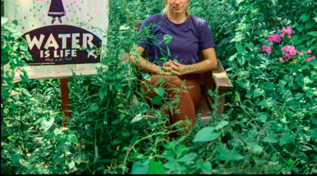 Photo by Cristina Yurena Zerr of Jessica Reznicek sitting among green plants and purple flowers next to a banner reading “We Are Here To Protect | Water Is Life”, other text reading “Support Jessica Reznicek & Navigating Conflict In Movement | TFSR 22-06-11”