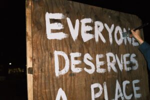 Hand painting "Everyone Deserves A Place" on a board with white paint. Photo by Elliot Patterson of Asheville Free Press